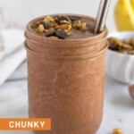 Chunky monkey smoothie in a glass jar with a straw in it. It is topped with chocolate chips and walnuts. Behind the jar are some bananas, a bowl of walnuts, and a spoon of cacao powder.