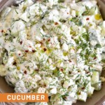 Cucumber yogurt salad garnished with fresh dill, olive oil, and red pepper flakes in a brown bowl. The bowel is still on a striped towel and next to it is half a lemon and a small dish of fresh dill.