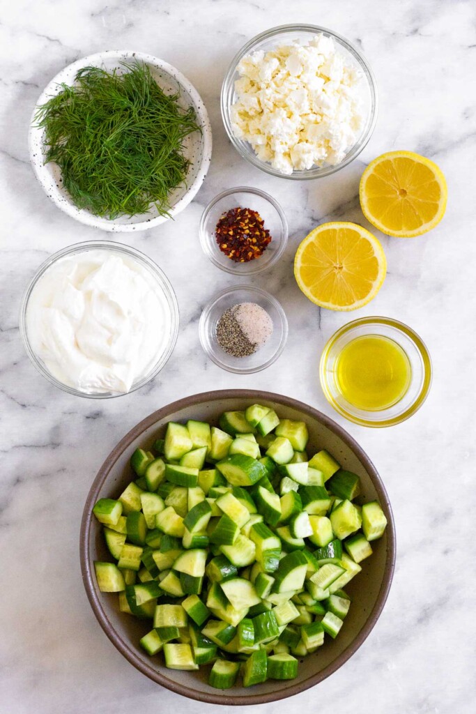 White marble counter with a bowl of feta cheese, a lemon cut in half, a bowl of olive oil, a bowl of chopped cucumbers, a bowl of salt and pepper, a bowl of red pepper flakes, a bowl of greek yogurt, and a plate of fresh dill.