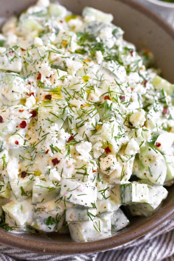 A close up of a large bowl of creamy cucumber dill salad garnished with fresh dill, red pepper flakes, and olive oil.