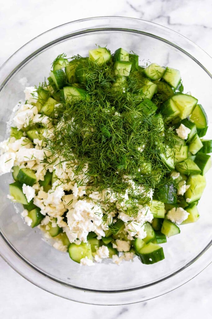 A glass bowl filled with a greek yogurt mixture with diced cucumbers, feta cheese, and fresh dill sitting on top of it before it is mixed together.