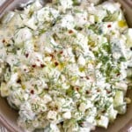 Cucumber yogurt salad garnished with fresh dill, olive oil, and red pepper flakes in a brown bowl. The bowel is still on a striped towel and next to it is half a lemon and a small dish of fresh dill.