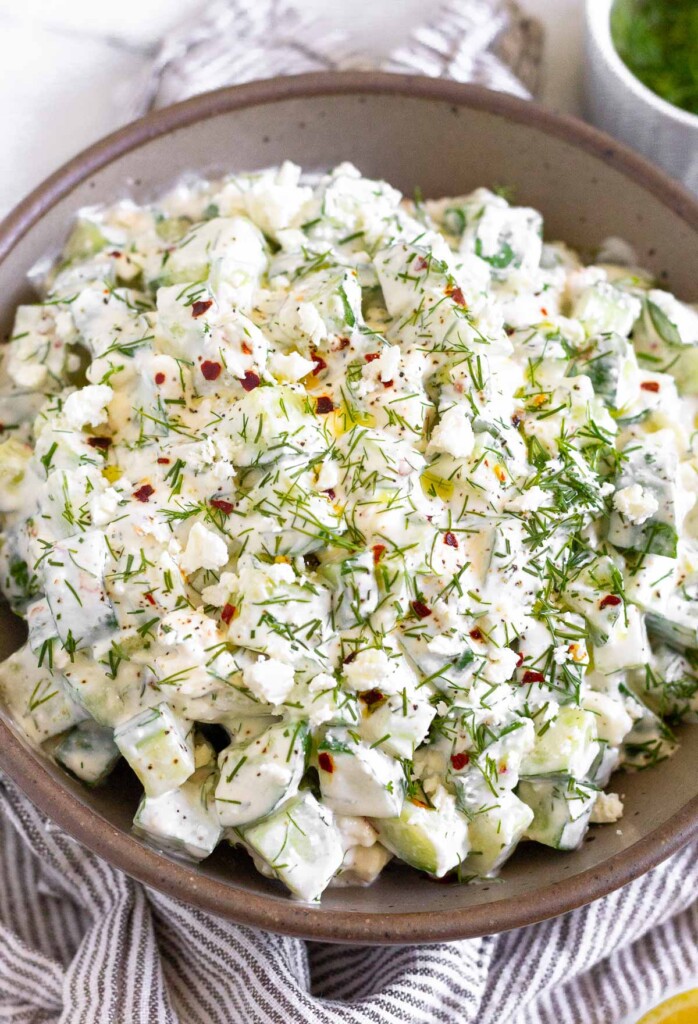 Cucumber feta salad in a large brown bowl garnished with fresh dill and red pepper flakes. The bowl is on a striped towel and around it is a half a lemon and a small dish of fresh dill.