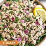 Italian tuna salad with capers in a bowl with arugula underneath it. It is garnished with 2 lemon wedges. Around the bowl is a half of lemon and 2 slices of bread.