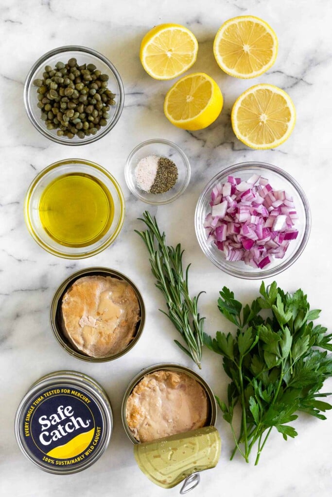 White marble counter with two lemons cut in half, a bowl of diced red onion, a small bowl of salt and pepper, a few sprigs of fresh rosemary, a bunch of fresh parsley, 2 cans of open tuna fish, 2 cans of tuna fish stacked on top of each other, a bowl of olive oil, and a bowl of capers.
