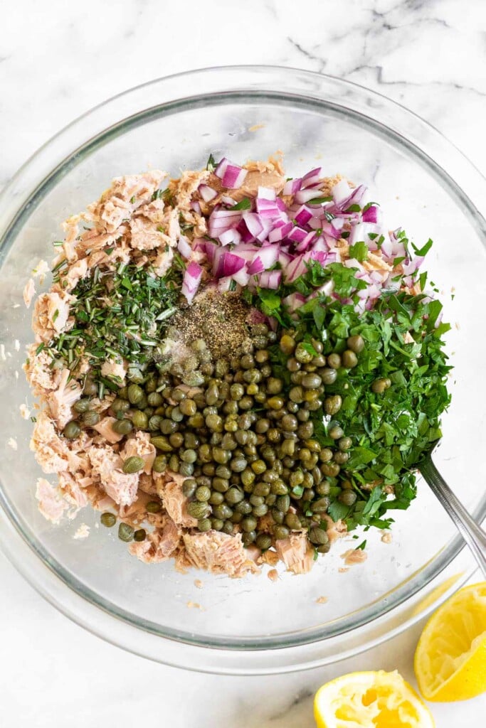 Large glass bowl with tuna fish, diced red onion, fresh parsley, capers, fresh rosemary, salt, and pepper before it is mixed together. Next to the bowl are two halves of juiced lemons.