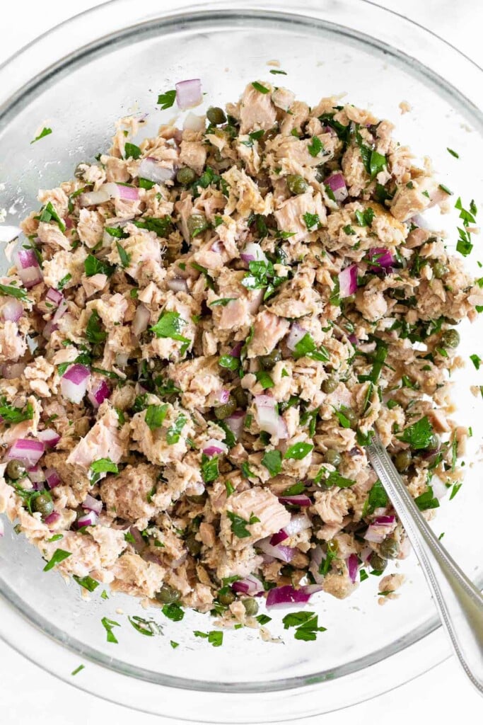 A large glass bowl of tuna fish mixed with red onion, fresh herbs, and capers with a fork in it.