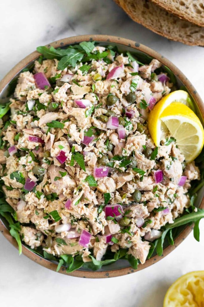 Tuna salad with capers, red onions, and fresh herbs overtop arugula in a bowl. It is garnished with two lemon wedges. Next to the bowl is half a juiced lemon and two slices of bread.