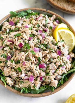 Italian tuna salad with capers in a bowl with arugula underneath it. It is garnished with 2 lemon wedges. Around the bowl is a half of lemon and 2 slices of bread.