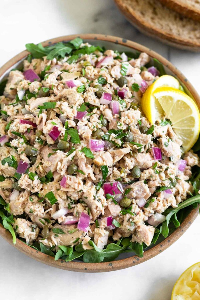 Italian tuna salad with capers in a bowl with arugula underneath it. It is garnished with 2 lemon wedges. Around the bowl is a half of lemon and 2 slices of bread.