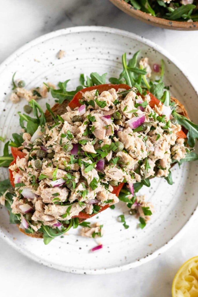 Overhead shot of tuna salad with olive oil, capers, red onion, and fresh herbs on top of sliced tomato and arugula on a piece of toast on a plate. Next to plate is half a juiced lemon a bowl of arugula.