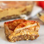 Close up of an apple peanut butter bar sitting on a white counter. Behind it is a pan of more bars, a spoon with peanut butter on it, and an apple.