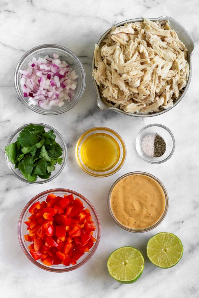 White marble counter with a bowl of shredded chicken, a small bowl of salt and pepper, a bowl of spicy mayo, a lime cut in half, a bowl of diced red bell pepper, a bowl of honey, a bowl of cilantro, and a bowl of diced red onion.