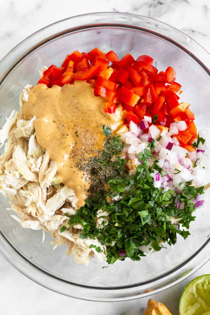 A large glass bowl of shredded chicken, diced red bell pepper, diced red onion, cilantro, chipotle mayo, honey, salt, and pepper before it is mixed together. Next to the bowl is half a juiced lime and a wooden reamer.