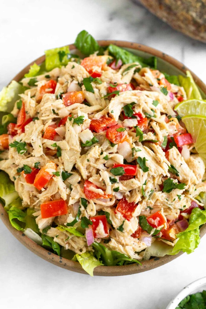 Chipotle chicken salad on top of some romaine lettuce in a bowl. It is garnished with lime wedges. Next to the bowl is a dish a cilantro and a loaf of bread.