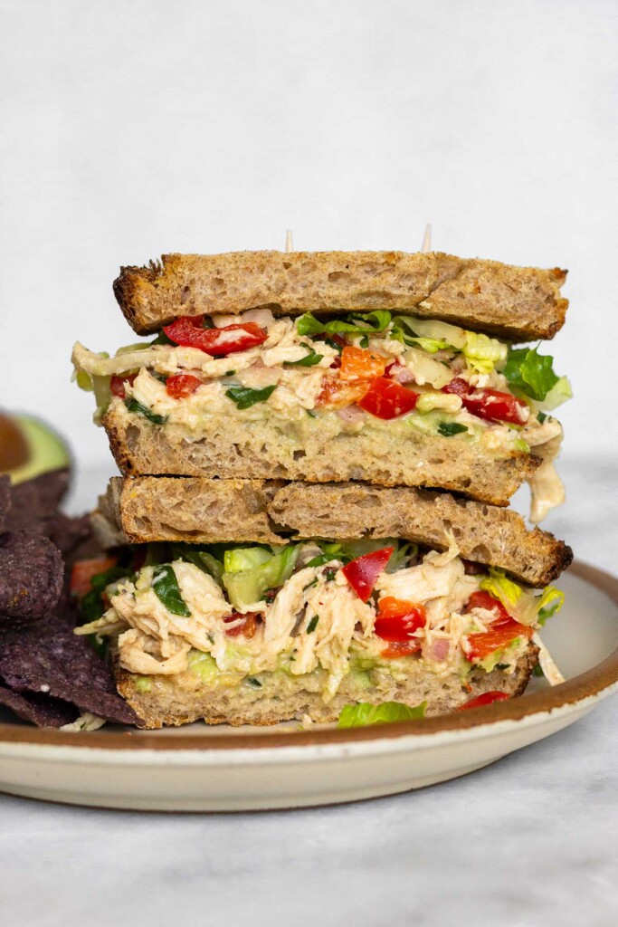 Two sandwich halves stacked on top of each other with lettuce, chicken salad, and avocado in them. There are on plate with some chip. Half an avocado is off in the background.