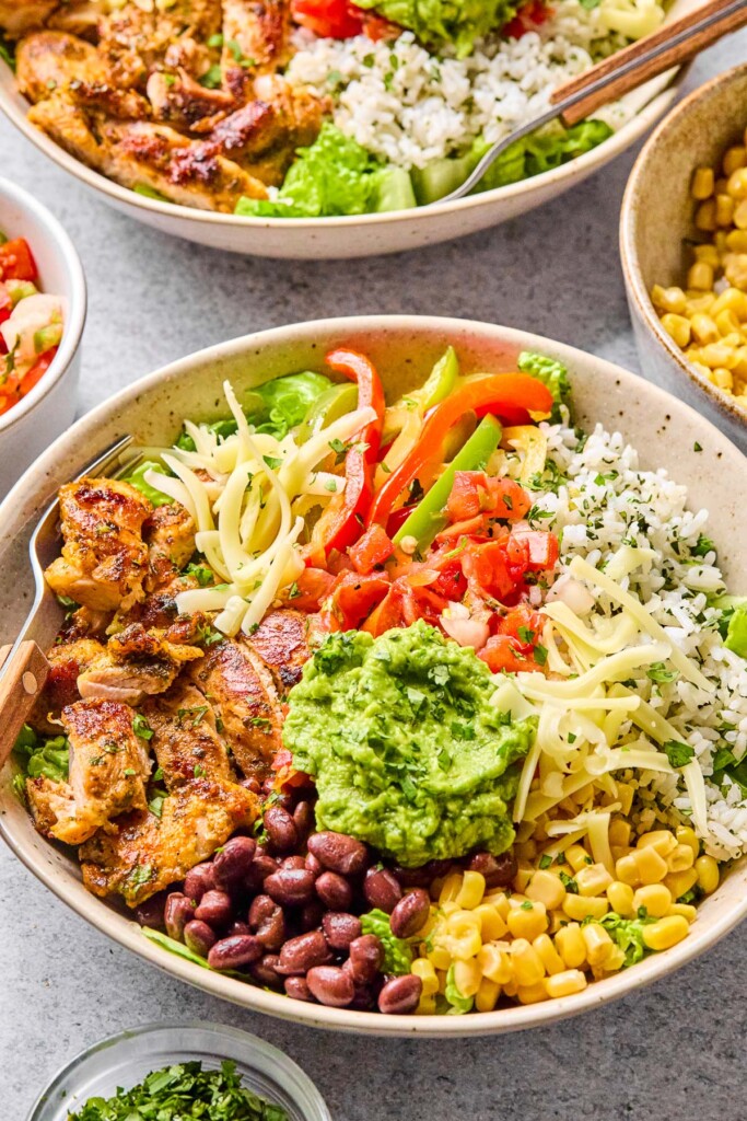 High protein meal prep bowl filled with seasoned chicken, fajita veggies, cilantro lime rice, black beans, corn, cheese, salsa, and guacamole. Around the bowl is a small bowl of cilantro, a bowl of salsa, another burrito bowl, and a bowl of corn.