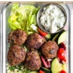 A high protein meal prep bowl filled with meatballs, cucumber and tomato salad, cauliflower rice, chopped romaine lettuce, and a small dish of tzatziki.