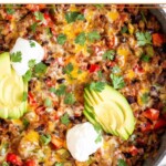 Taco skillet topped with melted cheese, sliced avocado, sour cream, and cilantro in a large sauté pan. Next to the skillet is a bowl of cilantro and two lime wedges.