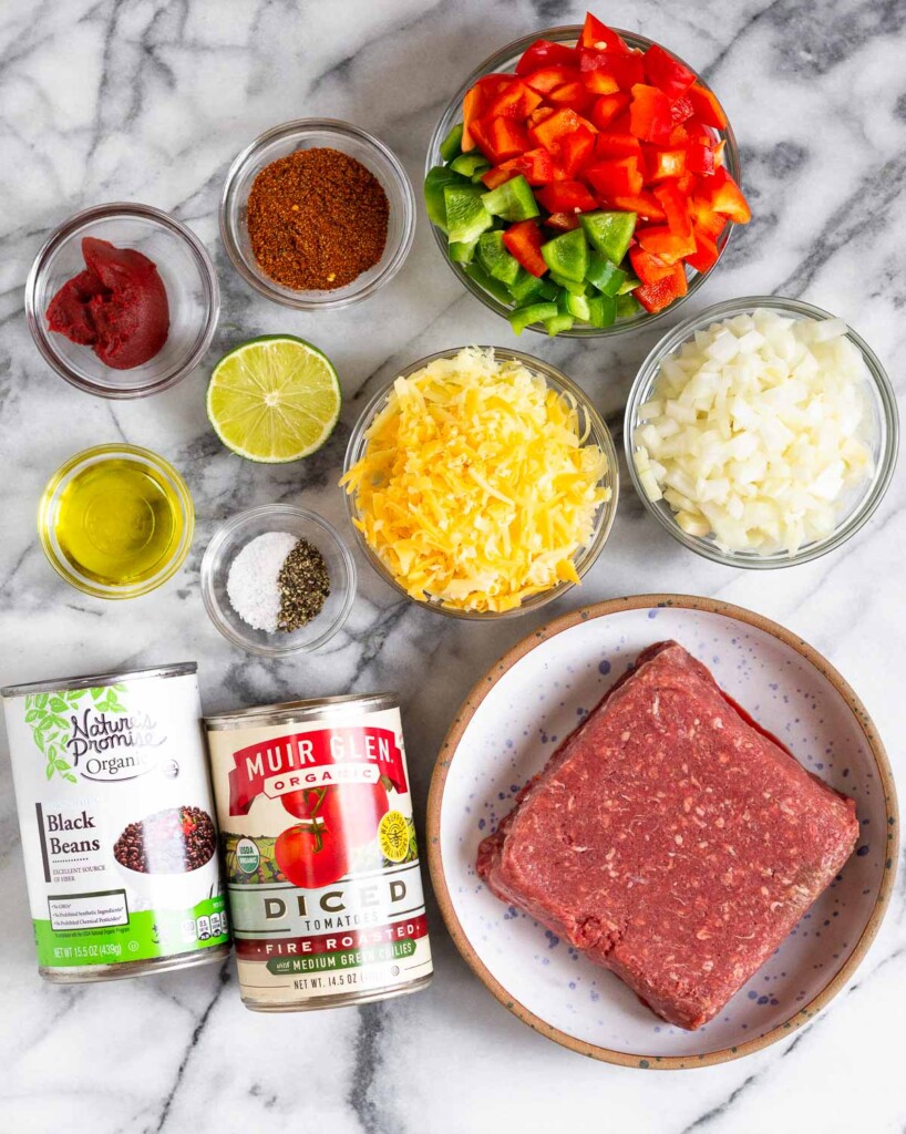 White marble counter with a bowl of diced red and green bell peppers, diced onion, a bowl of raw ground beef, a can of fire-roasted tomatoes, a can of black beans, a small bowl of salt and pepper, a bowl of shredded cheese, half a lime, a bowl of olive oil, a bowl of tomato paste, and a bowl of taco seasoning.