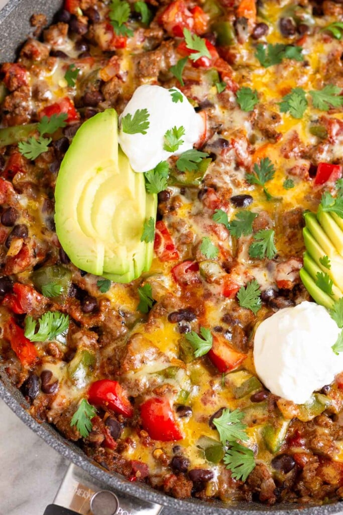 A skillet filled with cheesy taco skillet with ground beef, veggies, and black beans covered in melted cheese in a sauté pan. It is garnished with sliced avocado, sour cream, and cilantro.