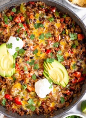 Taco skillet topped with melted cheese, sliced avocado, sour cream, and cilantro in a large sauté pan. Next to the skillet is a bowl of cilantro, two lime wedges, and a bowl of tortilla chips.
