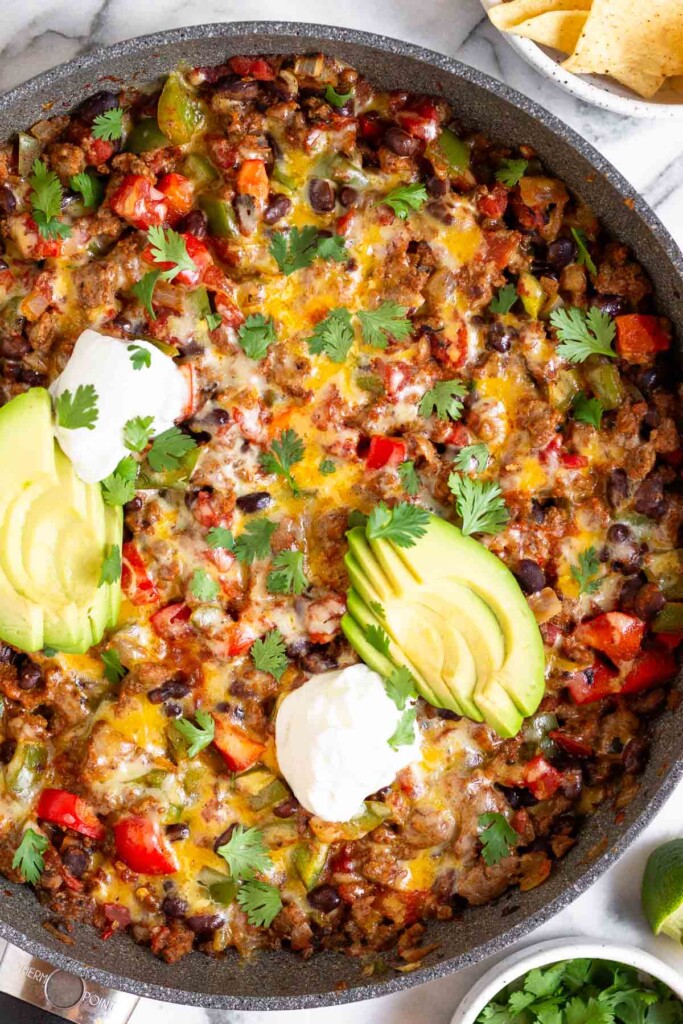 Taco skillet topped with melted cheese, sliced avocado, sour cream, and cilantro in a large sauté pan. Next to the skillet is a bowl of cilantro, two lime wedges, and a bowl of tortilla chips.