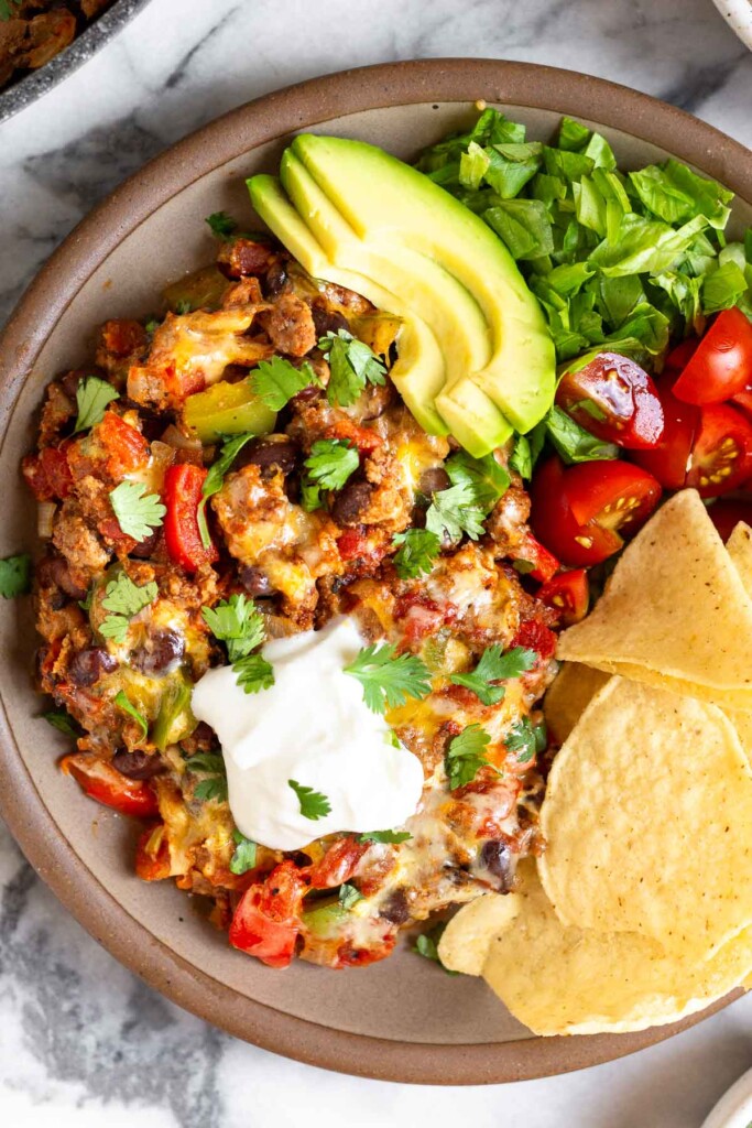A plate with mixture of ground beef, black beans, and veggies topped with melted cheese topped with sour cream and cilantro. Next to it are sliced avocado, chopped romaine, diced tomatoes, and tortilla chips.