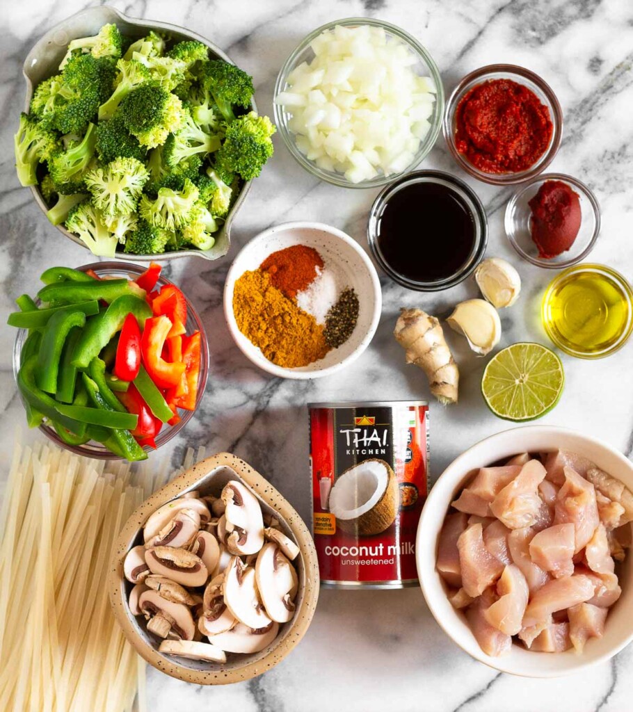 White marble counter with a bowl of diced onion, a bowl of red curry paste, a bowl of tomato paste, 2 garlic cloves, a bowl of olive oil, half a lime, a knob of ginger, a bowl of raw diced chicken, a can of full-fat coconut milk, a bowl of sliced mushrooms, a stack of uncooked rice noodles, a bowl of sliced bell peppers, a bowl of spices, and a bowl of broccoli florets.