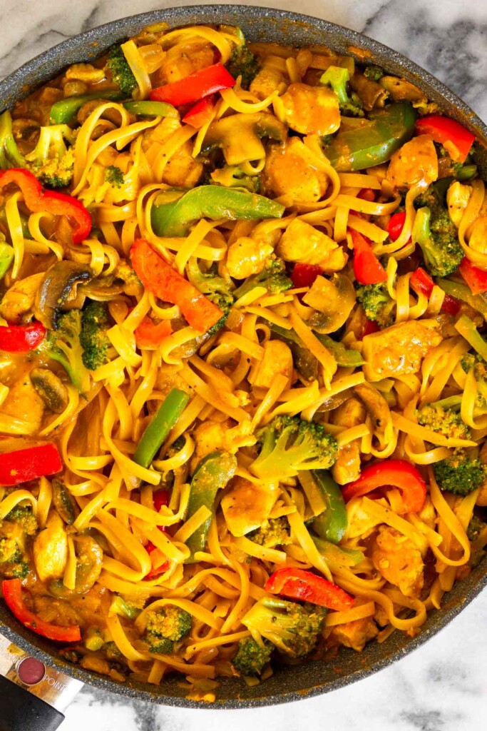 A skillet filled with noodles, chicken, broccoli, peppers, and mushrooms all tossed in red curry sauce.