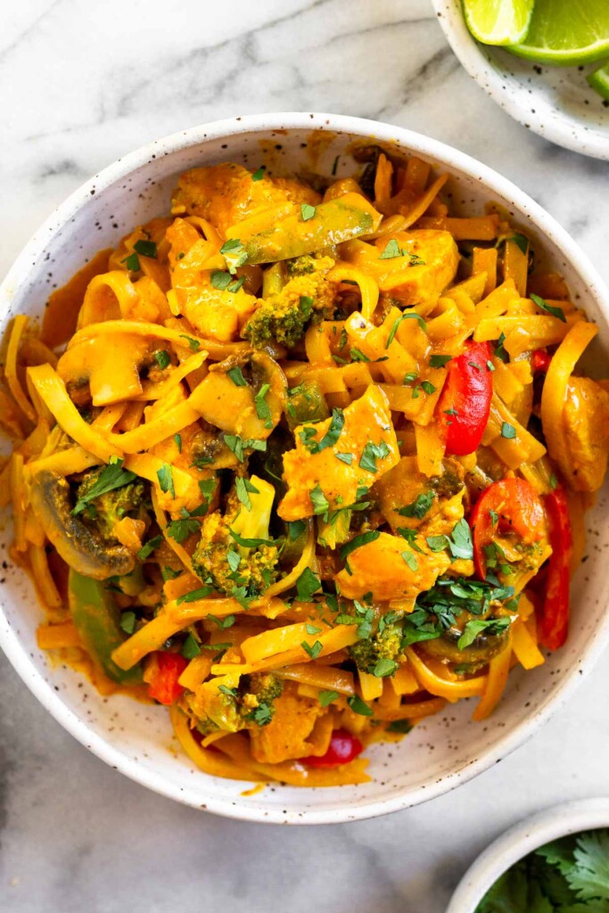 Red curry noodles with chicken, peppers, broccoli, and mushrooms in a white bowl. It is garnished with fresh cilantro. Next to the bowl is a plate of lime wedges and a bowl of cilantro.