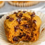 Close up of a gluten free pumpkin muffin with chocolate chips with a bite taken out of it. It is sitting on the muffin wrapper. Behind it is a muffin tin filled with more muffins.