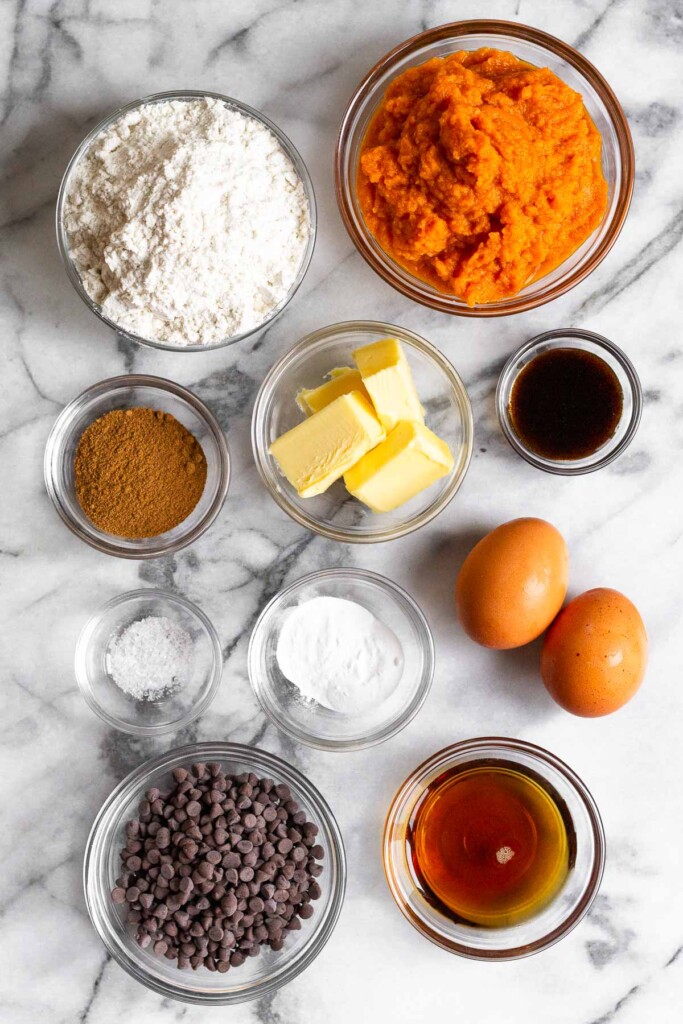 White marble counter with a bowl of pumpkin puree, a bowl of vanilla bean paste, 2 brown eggs, a bowl of maple syrup, a bowl of mini chocolate chips, a bowl of baking soda and powder, a bowl of salt, a bowl of cubed butter, a bowl of spices, and a bowl of flour.