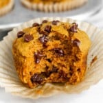 Close up of a gluten free pumpkin muffin with chocolate chips with a bite taken out of it. It is sitting on the muffin wrapper. Behind it is a muffin tin filled with more muffins.