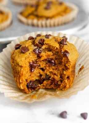 Close up of a gluten free pumpkin muffin with chocolate chips with a bite taken out of it. It is sitting on the muffin wrapper. Behind it is a muffin tin filled with more muffins.