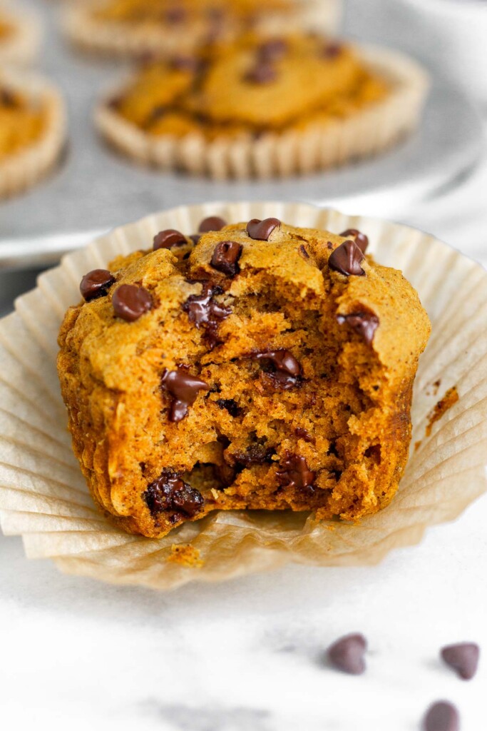Close up of a gluten free pumpkin muffin with chocolate chips with a bite taken out of it. It is sitting on the muffin wrapper. Behind it is a muffin tin filled with more muffins.