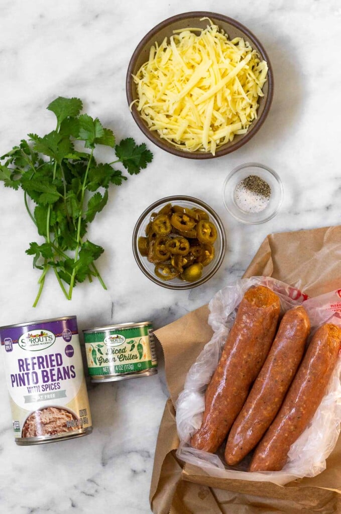 White marble counter with a bowl of shredded cheese, a small bowl of salt and pepper, a bowl of pickled jalapeños, 3 large uncooked sausage links, a can of green chiles, a can of refried beans, and a bunch of cilantro.