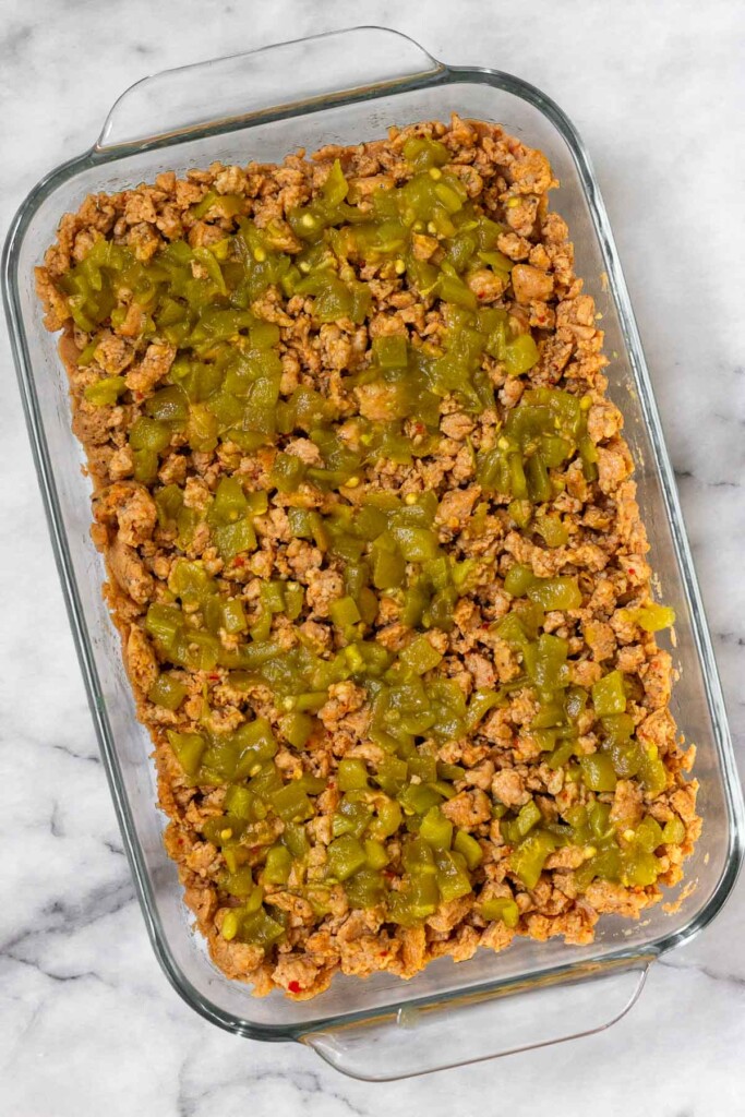 Overhead shot of a glass baking dish with refried beans evenly spread across the bottom topped with ground sausage and green chiles.