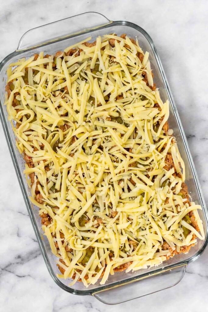 Overhead shot of a glass baking dish with shredded cheese overtop green chiles, ground meat, and beans before it is cooked.