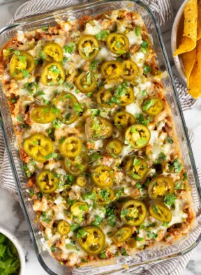 Cheesy sausage dip in a pan topped with pickled jalapeños and cilantro. The pan is on a striped towel and next to the pan is a bowl of tortilla chips and a bowl of cilantro.