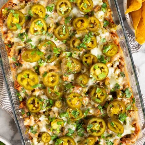 Cheesy sausage dip in a pan topped with pickled jalapeños and cilantro. The pan is on a striped towel and next to the pan is a bowl of tortilla chips and a bowl of cilantro.