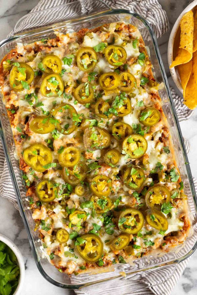 Cheesy sausage dip in a pan topped with pickled jalapeños and cilantro. The pan is on a striped towel and next to the pan is a bowl of tortilla chips and a bowl of cilantro.