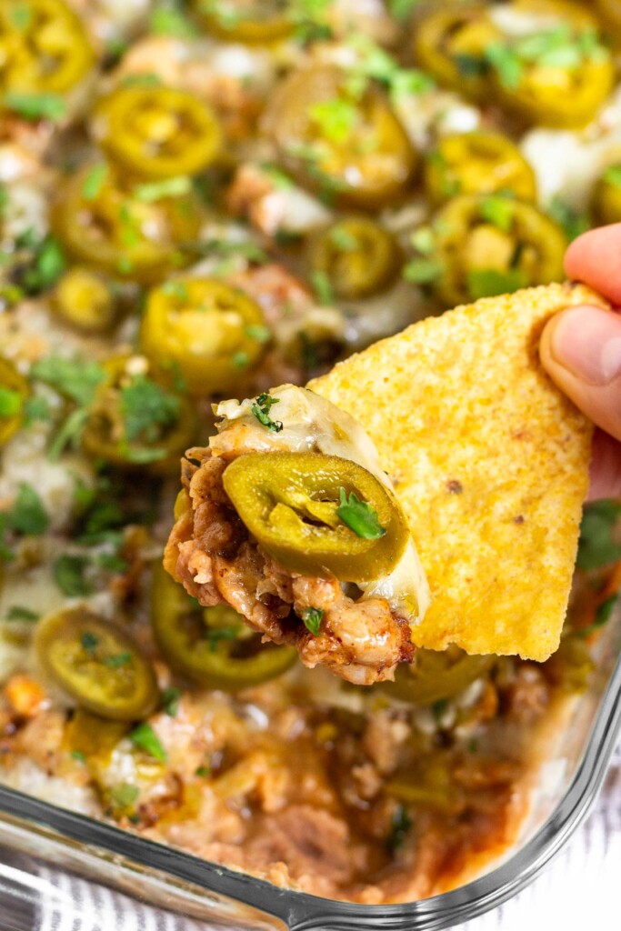 A hand holding a tortilla chip with hot sausage dip with pickled jalapeños on it. Behind it is a baking dish with the rest of the dip.