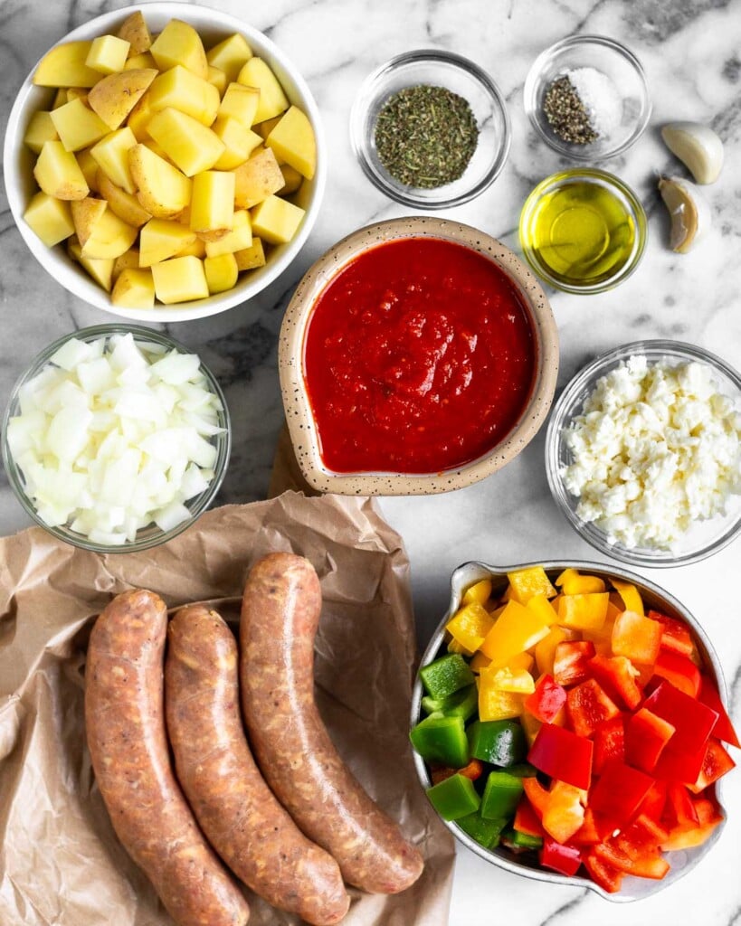 White marble counter with a small bowl of salt and pepper, a bowl of spices, a bowl of olive oil, two garlic cloves, a bowl of marinara sauce, a bowl of shredded cheese, a bowl of diced red, green, and yellow bell peppers, three large uncooked sausage links, a bowl of diced onions, and a bowl of diced potatoes.
