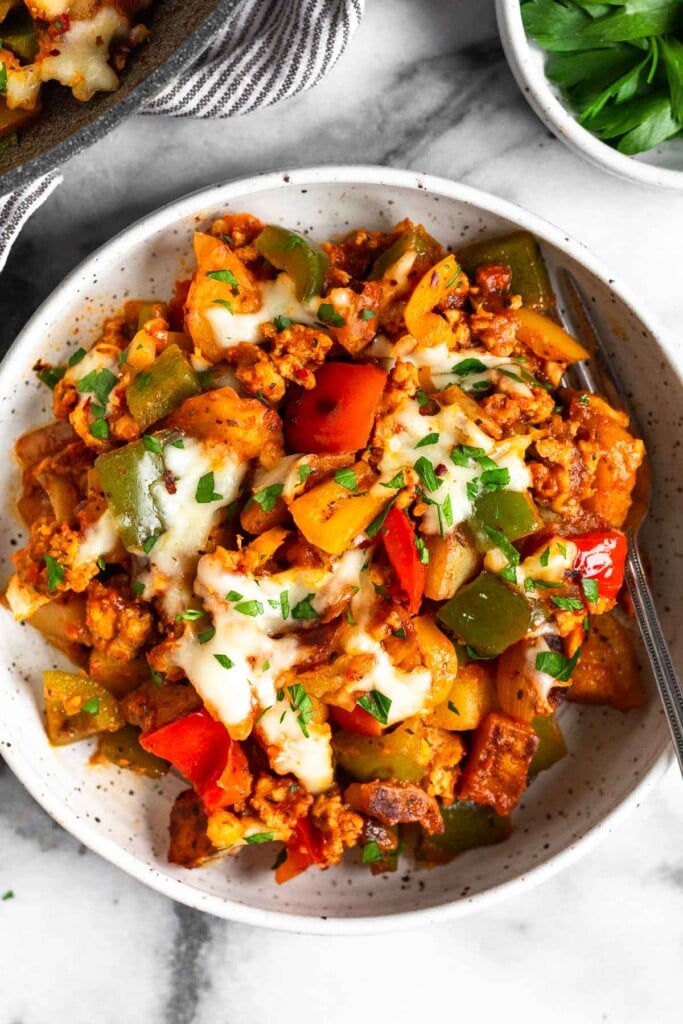 A bowl of sausage, potatoes, and peppers topped with melted cheese and garnished with fresh parsley. A fork is also in the bowl. Next to the bowl is a pan of more of the sausage mixture and a bowl of fresh parsley.