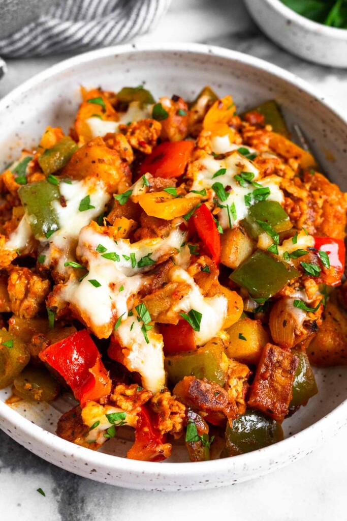 A bowl of a mixture of sausage with potatoes, peppers, and onions in marinara sauce topped with melted cheese. It is garnished with chopped herbs. Behind the bowl is a striped towel and a bowl of parsley.