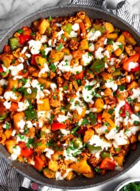 Sausage peppers and potatoes skillet topped with melted cheese and fresh parsley. The skillet is sitting on a striped towel and next to it is a small bowl of parsley.