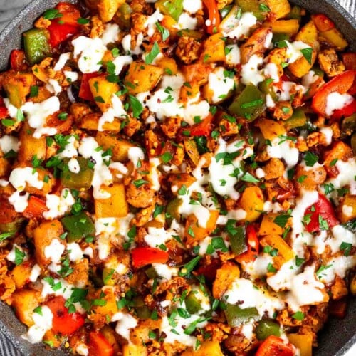 Sausage peppers and potatoes skillet topped with melted cheese and fresh parsley. The skillet is sitting on a striped towel and next to it is a small bowl of parsley.