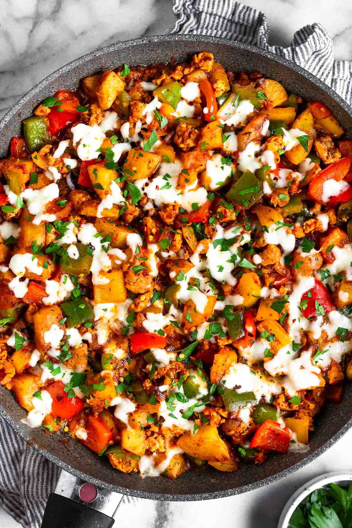 Sausage peppers and potatoes skillet topped with melted cheese and fresh parsley. The skillet is sitting on a striped towel and next to it is a small bowl of parsley.