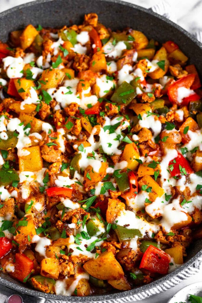 A close up of sausage potato skillet with peppers and onions. It is topped with melted cheese and garnished with fresh herbs.